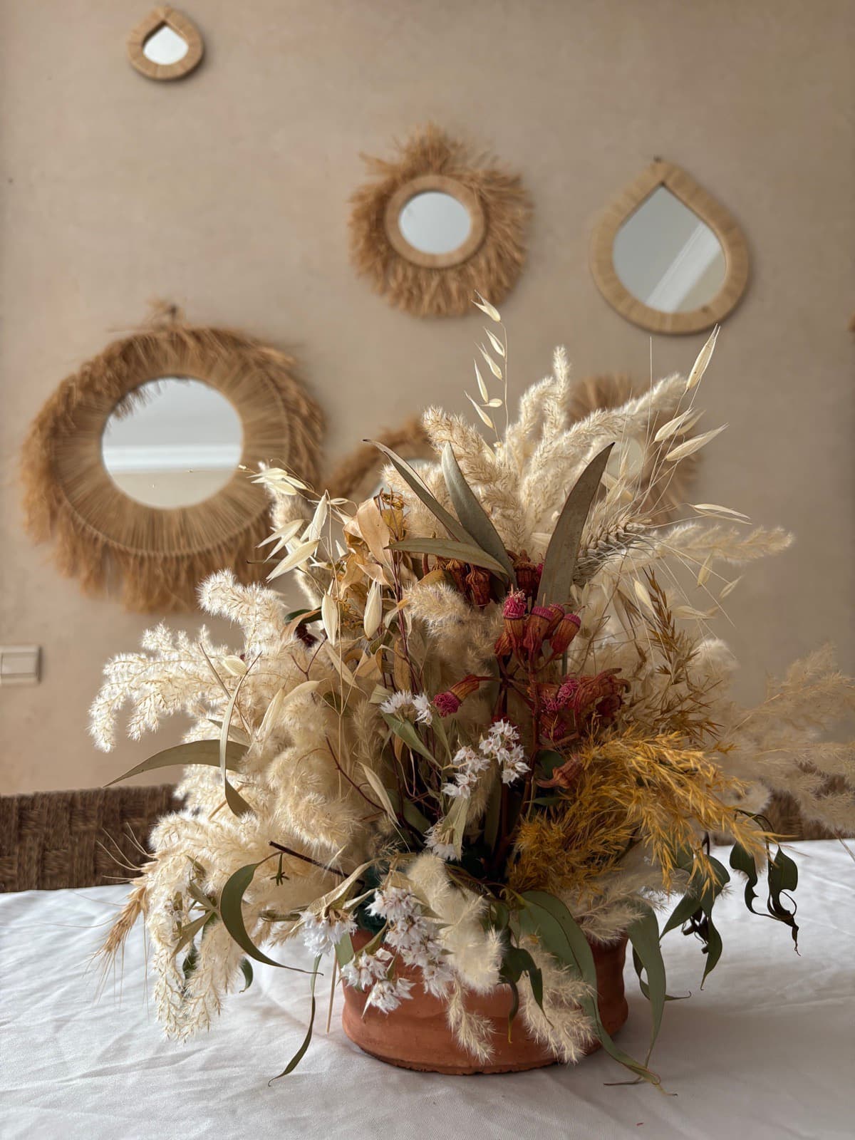 Bouquet de fleurs séchées dans un pot en terre cuite, devant un mur ocre orné de miroirs en raphia
